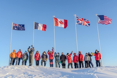 On a sunny day in the Arctic, 15 people bundled in cold-weather gear stand in a line with flags for Australia, France, Canada, United Kingdom, and USA waving on poles above them.