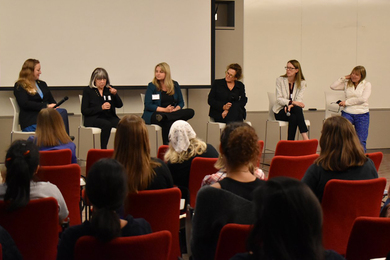 Six women, four of whom hold microphones, sit in a row facing an audience sitting in red chairs in a seminar room