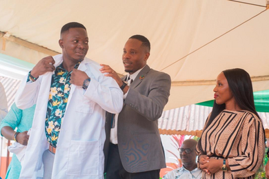 David Sengeh puts a white lab coat on Patrick Bangura. Claudine Humure looks on.