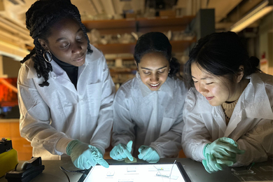 3 students in lab coats hunch over a table, looking at microfluidic devices 