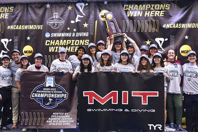 About 20 MIT student-athletes and coaches pose at a podium, hoisting their championship trophy overhead