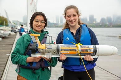 A pair of MIT students stand on a dock next to the Charles River holding a tube-like underwater vehicle