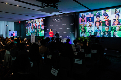 Hala Hanna speaks on a stage in a darkened room to a full audience. Two screens featuring many headshots flank her.