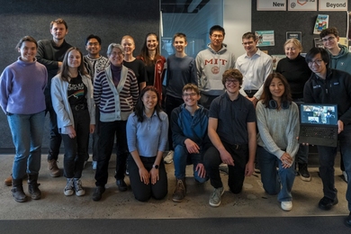 17 people pose in two rows for a group photo. One holds up a laptop screen to include half-a-dozen remote participants.