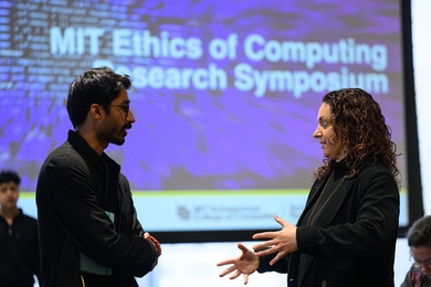 A man and a woman, seen in profile, converse in front of an MIT Ethics of Computing projection 