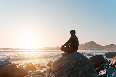 Silhouetted side view of a person wearing a hoodie sitting cross-legged on a rock overlooking the ocean.