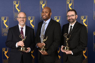 Joe McMaster, Wesley Richardson, and Clayton Hainsworth, all in suits, hold Emmy Award trophies in front of a wall of repeating Emmy logos that say “National Academy of Television Arts and Sciences Boston/New England Chapter.”