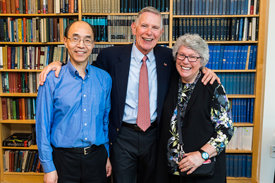 James Poitras stands in front of a large bookcase with his arms around the shoulders of Patricia Poitras and Guoping Feng