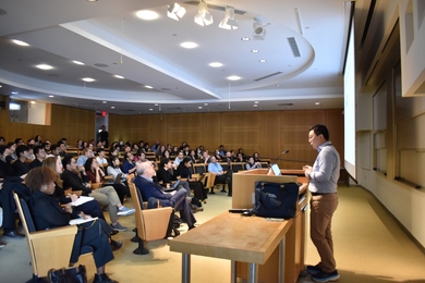 Side view of Li Ye, standing behind a lectern, speaking to a near-full audience at MIT's Singleton Auditorium