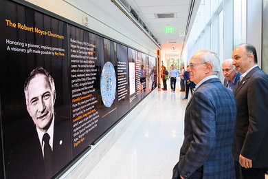 L Rafael Reif, Vladimir Bulović, and Ray Stata stand in a corridor looking at a wall with a picture of Robert Noyce and text on it.