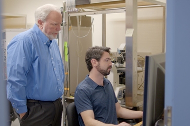 Andres Crane sits at a computer workstation in a lab. Troy Littleton stands behind him, looking at the screen