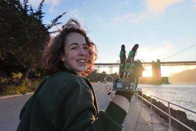 Freesia Gaul stands next to water and a large bridge, looking over her shoulder at the camera, holding up her hand wearing a haptic glove