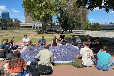 16 people sit around a large blanket in a grassy area on the Universidad Nacional Autónoma de México campus