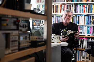 Joseph Paradiso sits at a desk in an office lined with bookshelves. He holds a sneaker fitted with electronic components and a small antenna.