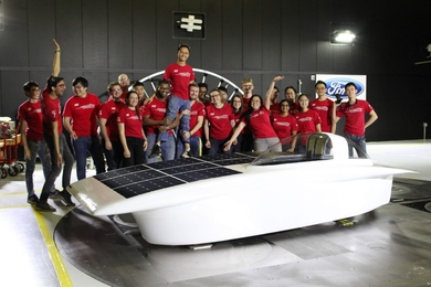 22 people in matching red T-shirts pose behind a solar electric vehicle. In the middle of the line, two men boost up Francis Wang