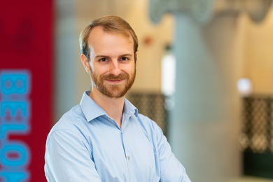 Dean Price poses indoors with a dark red banner and an architectural column in the background.