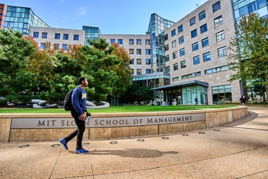 Student walks by the MIT Sloan School of Management sign in front of building E62 on a sunny day