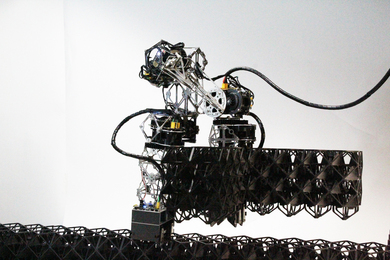 A robot, like an arm, holds 3 voxels. The voxels have a triangular lattice design and fit together.