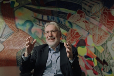 Duane Boning, seated in front of a colorful mural, gestures with both hands